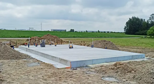 Płyta fundamentowa z wystającymi rurami na placu budowy, w tle pola i budynki.
